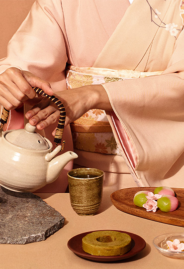 Person in a blush pink silk robe pouring tea from a ceramic teapot at a table set with Japanese-inspired desserts including matcha baumkuchen, green mochi, and cherry blossoms