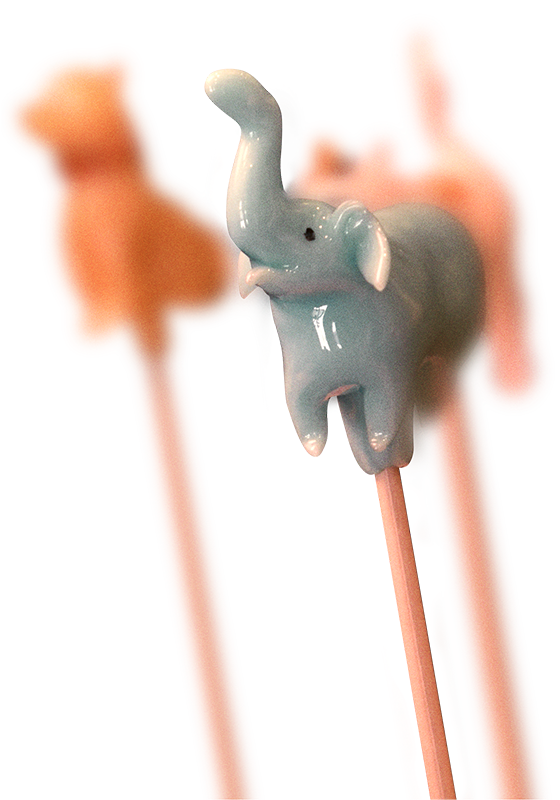 Light blue ceramic elephant figurine perched on wooden chopsticks with its trunk raised upward, photographed against a white background with blurred orange shapes in the distance
